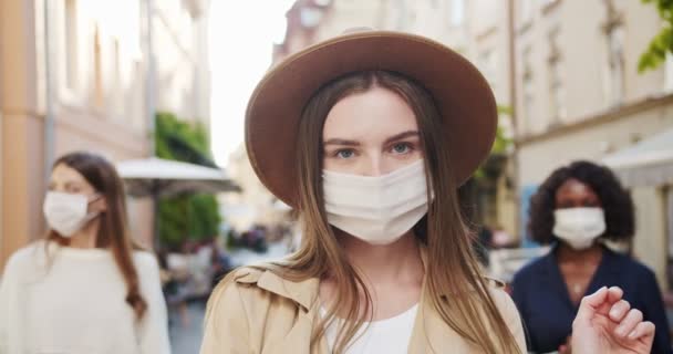 Primer plano retrato de la hermosa chica caucásica en sombrero y máscara médica de pie en la ciudad. Hermosa hembra afroamericana de fondo en máscara protectora. Cuarentena en la ciudad. Concepto de personas de raza mixta — Vídeos de Stock