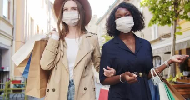 Piuttosto felici femmine di razze miste che camminano all'aperto con borse della spesa. Belle ragazze afroamericane e caucasiche in maschere mediche. Ragazza felice raggiunge i suoi amici per strada. Concetto di quarantena — Video Stock