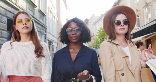 Neşeli çoklu etnik kadın portresi güneş gözlüğü takıyor ve alışverişlerle sokakta dururken gülümsüyor. Kameraya poz veren şapkalı neşeli beyaz kız. Güzel Afrikalı Amerikalı açık hava — Stok video