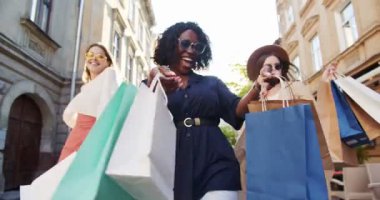 Güneş gözlüklü, alışveriş torbalarıyla sokakta yürüyen neşeli melez kızlar. Şehre taşınan mutlu beyaz güzel kızlar. Açık havadan alışveriş yapan Afro-Amerikalı bir kadın. Eğlenceli konsept