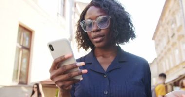 Güzel, Afro-Amerikalı genç bir kız alışveriş yaptıktan sonra şehirde dikilirken akıllı telefondan mesaj atıyor. Dışarıda cep telefonunda yazarken gülümseyen güzel bir kızın portresini yakından çek. Şehir konsepti