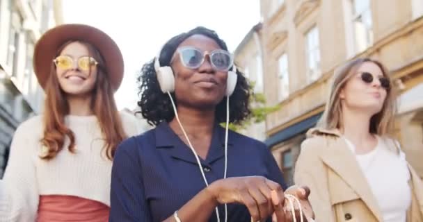 Hermosas mujeres alegres multi-étnicas caminando al aire libre después de ir de compras. Primer plano retrato de chica afroamericana escuchando música en auriculares en la calle.Feliz chicas caucásicas en gafas de sol sonriendo — Vídeo de stock