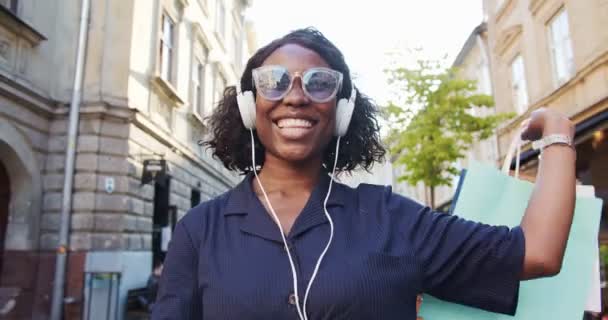 Feliz chica afroamericana divertida escuchando música en los auriculares mientras camina en la ciudad. Primer plano retrato de hermosa hembra en gafas de sol con compras después de hacer compras en la calle. Concepto divertido — Vídeos de Stock