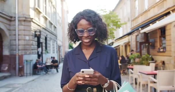 Primer plano retrato de alegre bonita afroamericana joven mensajes de texto en el teléfono inteligente mientras camina por la ciudad. Hermosa chica con bolsas de compras sonriendo mientras escribe en el teléfono celular al aire libre. Concepto de ciudad — Vídeos de Stock