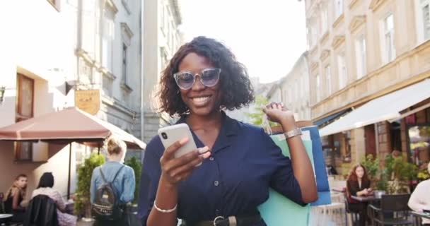 Alegre afroamericana joven mujer mensajes de texto en el teléfono inteligente mientras camina en la ciudad después de ir de compras. Primer plano retrato de hermosa chica feliz con compras sonriendo mientras se toca en el teléfono celular al aire libre — Vídeos de Stock