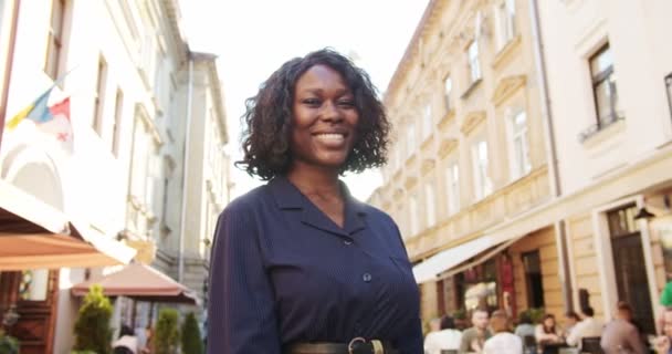 Primer plano retrato de hermosa chica alegre sonriendo a la cámara mientras que de pie al aire libre y sosteniendo bolsas de compras. Mujer afroamericana alegre con sonrisa en la cara de pie en la calle. Concepto de emociones — Vídeos de Stock