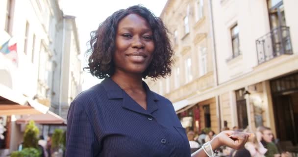 Primer plano retrato de hermosa chica alegre sonriendo a la cámara mientras está de pie al aire libre. Alegre bonita mujer afroamericana en blusa azul con sonrisa en la cara de pie en la calle. Concepto de emociones — Vídeos de Stock