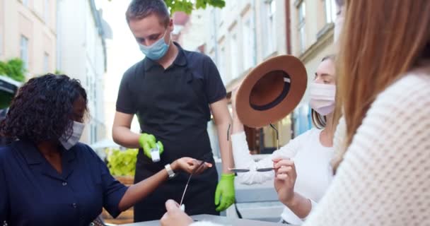 Olika blandras flickor i masker sitter vid bordet i cafeteria terrass. Manlig servitör mäter temperaturen hos vackra afroamerikanska och kaukasiska flickor. Stadsliv i karantän. Utomhuskoncept — Stockvideo