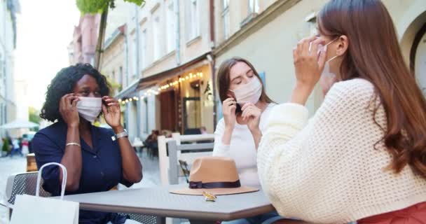 Belles femmes multi-ethniques assises à la cafétéria à l'extérieur. Joyeuses filles afro-américaines et caucasiennes repoussant les masques médicaux et ayant une conversation. Communication en quarantaine. Concept de ville — Video