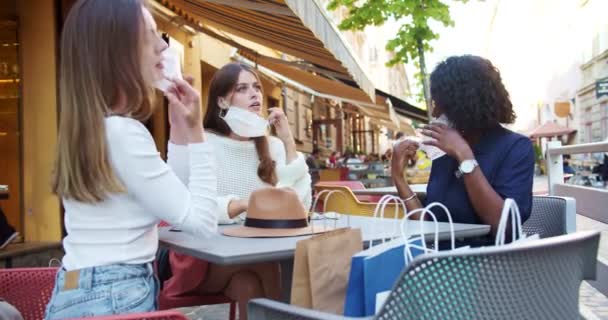 Smíšené ženy sedící na terase restaurace a odkládající lékařské masky. Krásné africké Američanky a bělošky po nakupování sedí u stolu venku s úsměvy na tvářích. Koncept města — Stock video