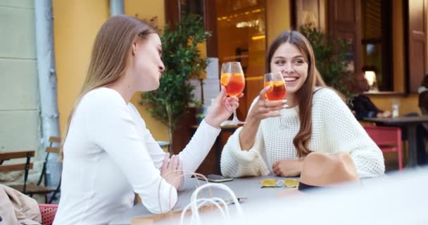 Dos hermosas niñas caucásicas sentadas en la mesa de la cafetería al aire libre y bebiendo cócteles de verano. Bastante alegre hembras charlando mientras bebe bebidas en la ciudad. Estilo de vida urbano. Concepto de ocio — Vídeo de stock