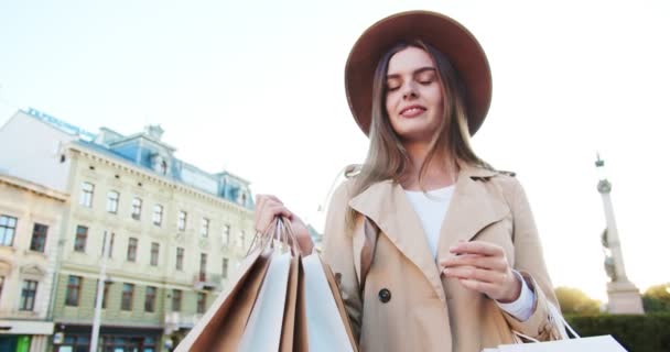 Portræt af glad smuk pige i hat og frakke holder poser mens stående udendørs i byen. Smuk hvid kvinde i godt humør smilende efter shopping på gaden. Shopper koncept – Stock-video