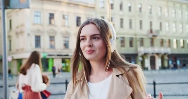 Primer plano retrato de la joven caucásica alegre en la calle escuchando música en los auriculares. Hermosa chica feliz bailando mientras camina en la ciudad y disfruta de canciones al aire libre. Concepto de ocio — Vídeo de stock