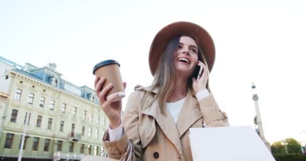 Menina feliz em chapéu e casaco com sacos de compras em pé ao ar livre e falando no smartphone enquanto segura xícara de café. Mulher branca bonita sorrindo enquanto conversa no celular na rua — Vídeo de Stock