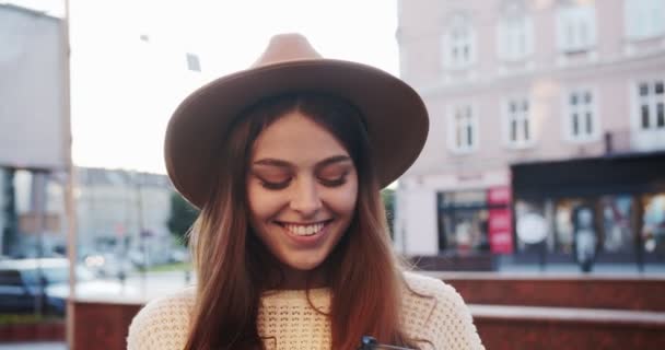Közelkép portré gyönyörű kaukázusi fiatal nő mosolyog a kamera, miközben áll az utcán a városban. Vidám, csinos lány kalapban, mosollyal az arcán, aki elnéz a szabadban. Érzelmek fogalma — Stock videók