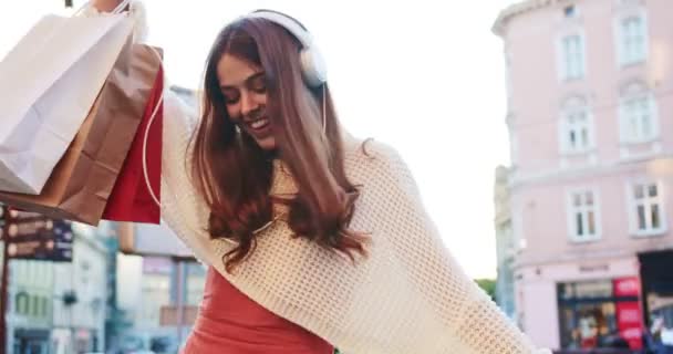 Jovem caucasiana feliz ouvindo música em fones de ouvido e dançando na rua na cidade. Retrato de menina bonita alegre se movendo e se divertindo enquanto está de pé com sacos de compras. Conceito de comprador — Vídeo de Stock