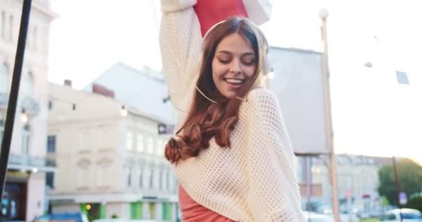 Porträtt av glad vit ung kvinna som lyssnar på musik i hörlurar och dansar på gatan. Vacker glad flicka rör sig stående med inköp efter shopping och njuta av sång. Kul koncept — Stockvideo