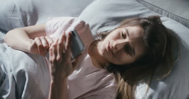 Blick von oben auf eine junge attraktive Frau aus dem Kaukasus, die frühmorgens unter einer Decke im Bett liegt und SMS auf ihr Smartphone sendet. Draufsicht auf hübsches lächelndes Mädchen beim Anzapfen des Telefons im Schlafzimmer. — Stockvideo