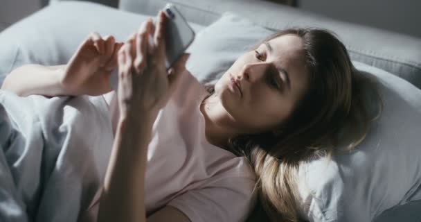 Верхний вид на кавказскую молодую красивую женщину, лежащую в постели рано утром и переписывающуюся на смартфоне. Вид сверху на красивую девушку, постукивающую и прокручивающую по мобильному телефону в спальне. — стоковое видео