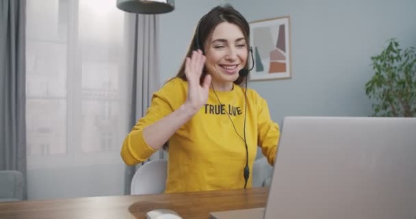 Jovem caucasiana bonita alegre acenando com a mão e falando em videocall no laptop em fone de ouvido em casa. Menina bonita feliz sorrindo enquanto vídeo conversando no computador no quarto. Conceito de lazer — Vídeo de Stock