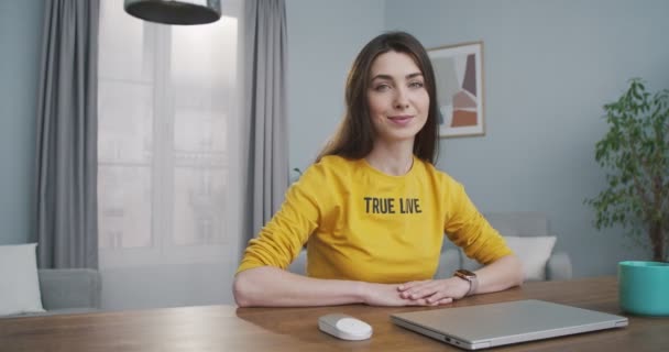 Bastante caucásico alegre joven hembra de buen humor sonriendo mientras mira a la cámara en la habitación acogedora. Chica hermosa alegre en suéter amarillo sentado en la mesa en casa con sonrisa en la cara. Concepto interior — Vídeo de stock