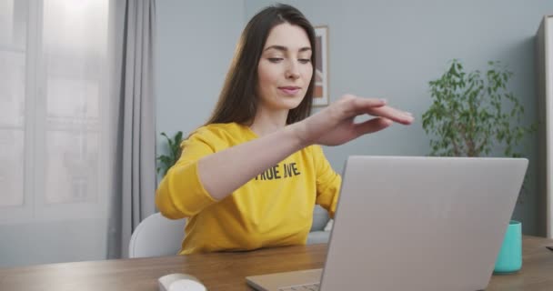 黄色のプルオーバーで幸せな美しい女の子は、コンピュータを開き、自宅で作業しながら入力します。居心地の良い部屋でテーブルに座っている間、かなり白人の喜びの若い女性がラップトップでブラウジング。フリーランスのコンセプト — ストック動画