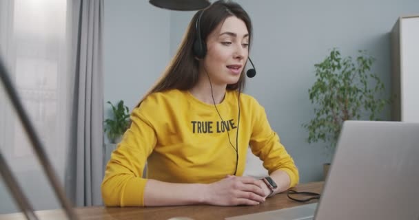 Nešťastná krásná kavkazská mladá žena, která má videokonverzaci na notebooku ve sluchátkách v pokoji doma. Hezká dívka video chatování na počítači, zatímco sedí u stolu uvnitř. Koncept videohovoru — Stock video
