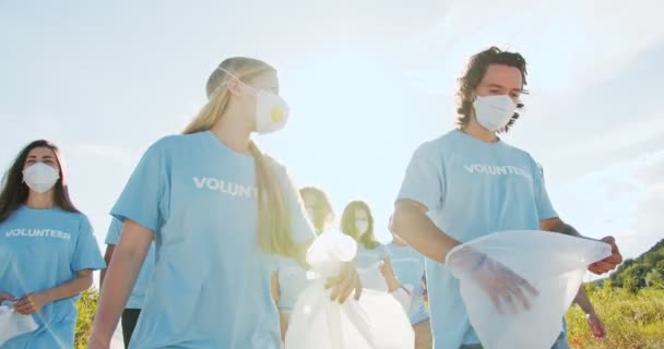 Un grupo de voluntarios yendo en máscaras médicas con basura, bolsas de basura para recoger botellas de plástico, residuos. Un líder de conservacionistas de cinco, animando e impulsando el espíritu de equipo. Voluntariado. — Vídeos de Stock