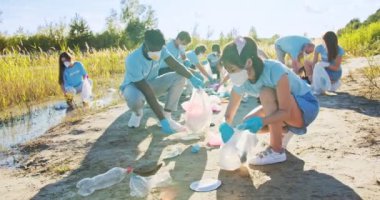 Kirlilikten plajı temizleyen çok ırklı tıbbi maskeli gönüllüler. Eldivenli çevreciler plastik şişeleri ve poşetleri topluyorlar. Çevre, ekoloji, biyosfer koruma kavramı.