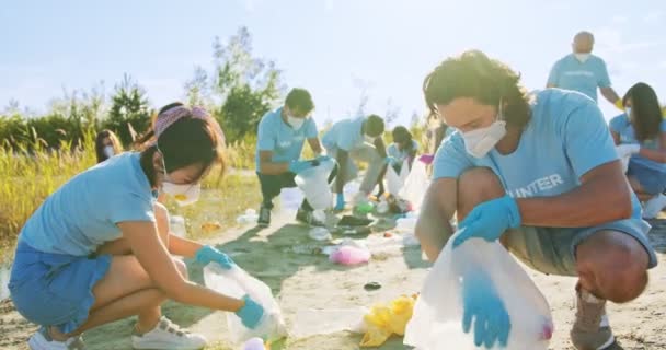 Activistas caucásicos masculinos y femeninos asiáticos en máscaras recogiendo botellas de plástico, bolsas y residuos en la playa. Voluntarios entusiastas recogiendo basura, basura. Concepto de conservación ambiental. — Vídeos de Stock