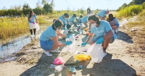 Hombres y mujeres voluntarios con máscaras médicas limpiando la playa de basura, basura. Activistas multiétnicos recogiendo artículos de plástico en una bolsa de basura. Salva la Tierra. Concepto de problema ecológico. — Vídeos de Stock