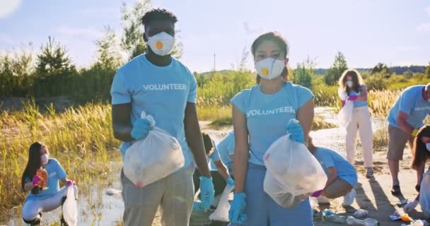 Le groupe de bénévoles en masques ramassant des ordures, des ordures près du rivage. Des écologistes multiethniques tenant des sacs poubelles et regardant la caméra. Volontariat, concept de travail d'équipe. — Video