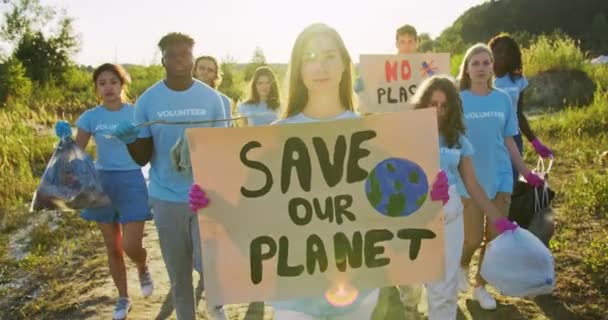 Una voluntaria caucásica protestando contra la contaminación del planeta. Un grupo de eco-acivistas multiétnicos caminando con la basura recogida y el cartel de Save Our Planet. Conservación ambiental. — Vídeo de stock