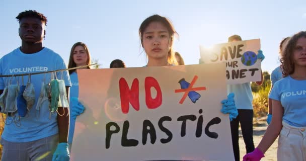 Organización voluntaria de eco activistas multiétnicos que protestan contra la utilización del plástico, la contaminación. Joven mujer asiática voluntaria caminando con cartel No Plastic y mirando a la cámara. Voluntariado. — Vídeos de Stock