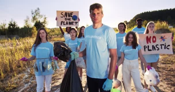 Група багатонаціональних екологічних захисників з сміття і плакатів "Бережіть нашу планету, без пластику". Нахабний кавказький доброволець, який тримає сміття, сміттєвий мішок і дивиться на камеру. Добровольча ідея. — стокове відео