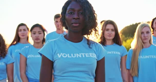 Jovem mulher atraente com um grupo de ativistas da natureza em segundo plano. Bonita voluntária africana olhando para a câmera e dizendo: Eu sou uma voluntária. Voluntariado, conservação do ambiente . — Vídeo de Stock