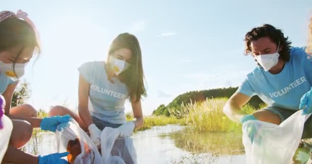 Activistas ecológicos limpiando el agua de la basura, basura. Un grupo de voluntarios en máscaras médicas y guantes recogiendo botellas de plástico, residuos, basura y poner en bolsas. Concepto de supervivencia ecológica. — Vídeos de Stock