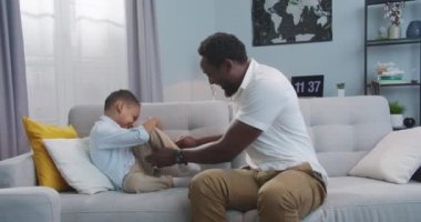 Yakışıklı, neşeli bir adam küçük oğluyla eğleniyor ve oturma odasında gülümsüyor. Neşeli Afrikalı Amerikalı baba evde, kanepede sevimli, mutlu küçük çocukla oynuyor. Aile kavramı