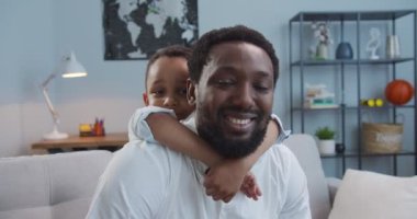 Yakışıklı, neşeli, Afro-Amerikan bir adamın küçük sevimli oğluyla oturma odasında gülümseyen portresi. Sevimli, neşeli bir çocuk. Evinde, kanepede babasına sarılıyor. Mutlu aile kavramı