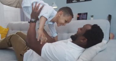 Neşeli küçük şirin çocuk kanepeye atlıyor ve babasıyla oynuyor. Yakışıklı, mutlu, Afro-Amerikan genç bir baba odada kanepede yatarken oğluyla uçak oyunu oynuyor. Aile kavramı