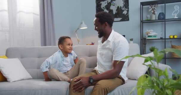 Bonito menino afro-americano feliz sentado em casa no sofá e abraçando seu pai dentro de casa. Bonito homem alegre com criança pequena sorrindo na sala de estar aconchegante de bom humor. Conceito familiar — Vídeo de Stock