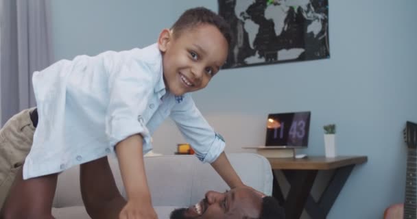 Feche o retrato do menino feliz bonito brincando com o pai em casa. Bonito homem afro-americano alegre levantando o pequeno filho como um avião voador enquanto estava deitado no sofá na sala de estar. Conceito familiar — Vídeo de Stock