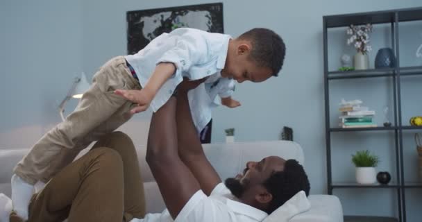 Knappe vrolijke Afro-Amerikaanse vader spelen vliegtuig spel met schattige zoon terwijl liggend op de bank in de kamer. Vreugdevolle man tilt kleine gelukkige zoon op alsof hij binnen vliegt. Vrijetijdsconcept — Stockvideo