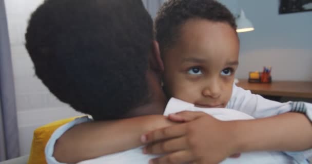 Close up retrato de uma criança pequena bonito abraçando seu pai afro-americano em casa. Um rapazinho simpático a abraçar o pai na sala de estar dentro de casa. Uma família adorável. Relações pai-filho. Conceito de infância — Vídeo de Stock