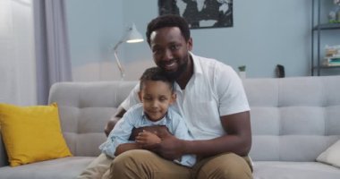 Evdeki kanepede küçük tatlı oğluyla otururken gülümseyen mutlu Afrikalı Amerikalı genç babanın portresi. Yakışıklı, neşeli bir adam. Oturma odasında küçük neşeli çocuğa sarılıyor. Aile kavramı