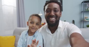 Mutlu Afro-Amerikan yakışıklı erkek ve küçük oğlu evde akıllı telefondan video sohbeti yaparken el sallıyor. Neşeli baba ve sevimli çocuk cep telefonuyla video görüşmesi yapıyorlar. Boş zaman konsepti