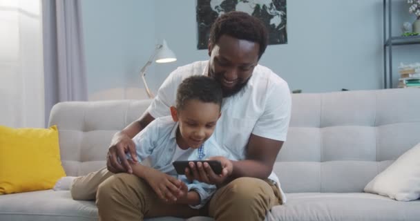 小さなかわいい息子と一緒にスマートフォンでビデオを見て幸せなアフリカ系アメリカ人男性と自宅でソファに座っている間に動きを模倣します。小さな子供に携帯電話で何かを示すハンサムな楽しい父親 — ストック動画
