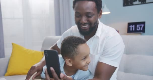 Portrait d'homme afro-américain heureux avec un petit fils mignon parlant sur appel vidéo sur smartphone à la maison. Beau père avec petit garçon ayant une vidéoconférence sur téléphone portable tout en étant assis Concept d'appel — Video