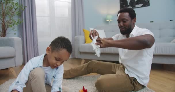 행복 한 아프리카계 미국인 소년이 바닥에 앉아 있는 동안 집 거실에서 아빠와 노는 모습. 잘 생기고 잘생긴 남자가 어린 아들과 재미있게 놀고 있어요. 아버지와 아이들이 장난감을 가지고 노는 모습 — 비디오