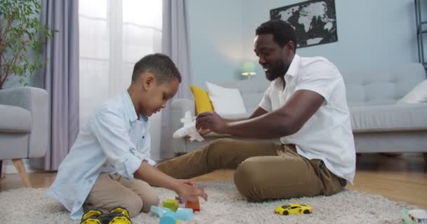 Веселий афроамериканець грає з батьком у вітальні, сидячи на підлозі. Гарний усміхнений чоловік розважається з гарним сином. Тато та дитина бавляться іграшками. Дивна концепція — стокове відео